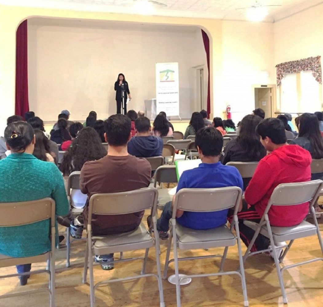 Coach Priya standing on a small stage at a microphone, addressing rows of seated children and parents beside the DSI banner