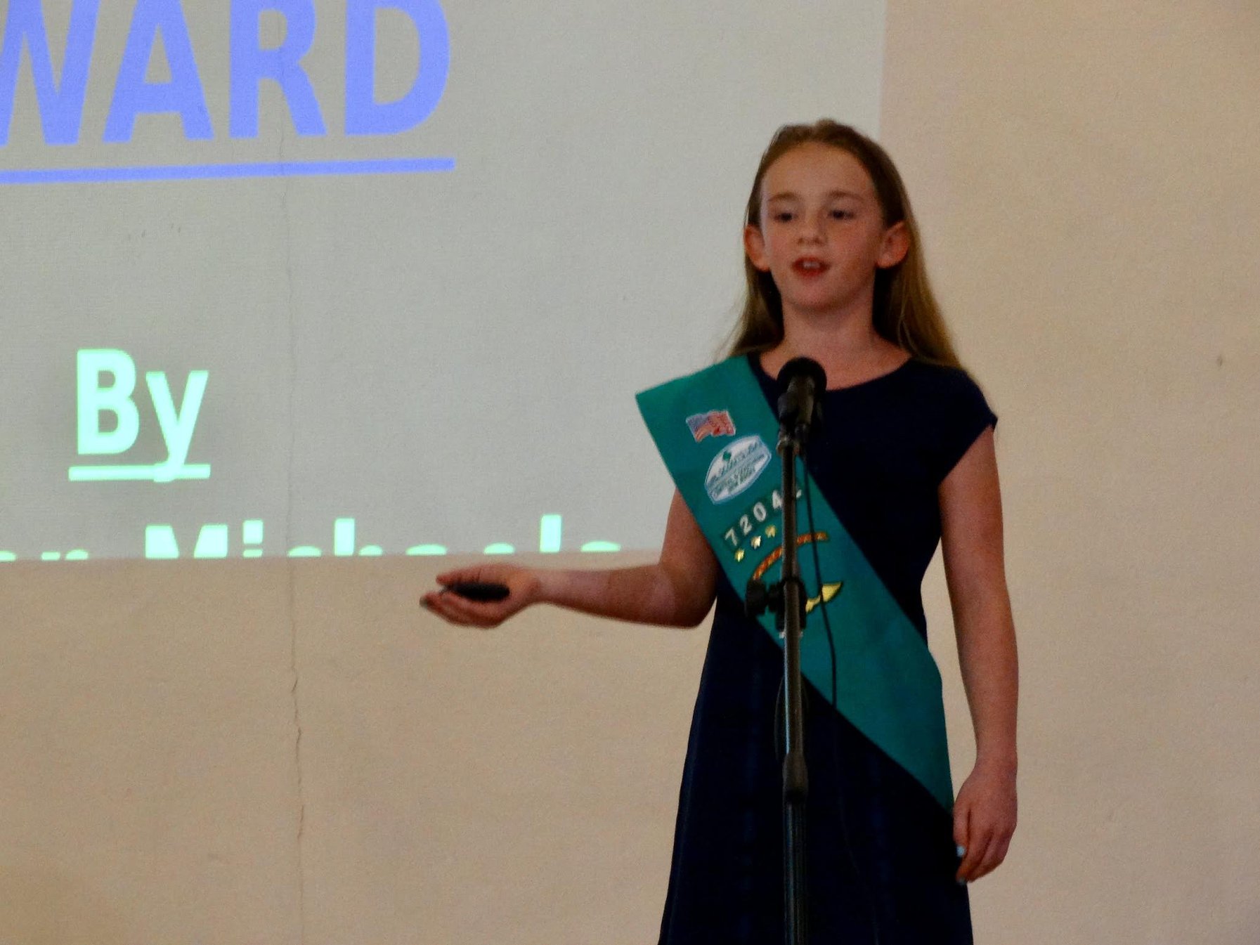 A young DSI speaker in a Girl Scouts sash presenting at the microphone beside a projected award slide