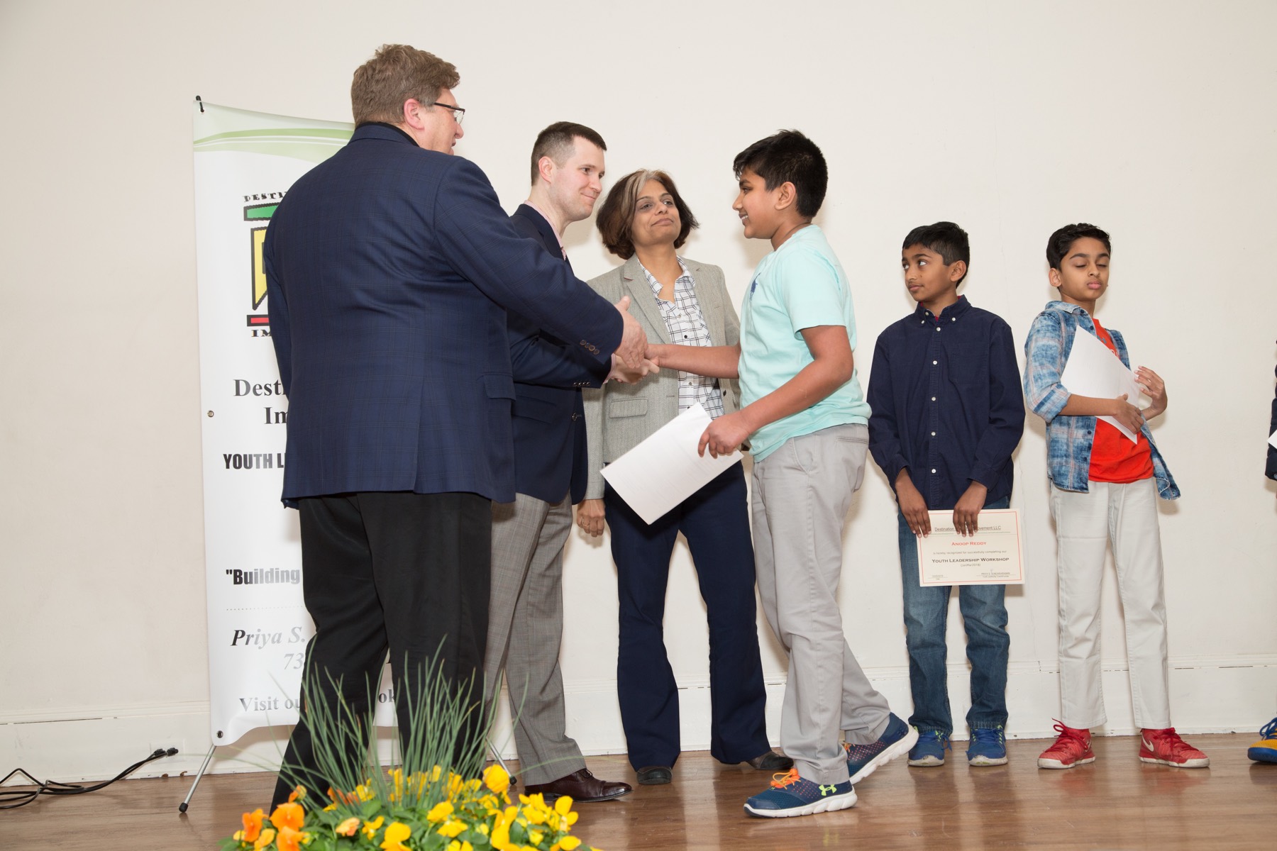 A DSI student shaking hands with a judge while receiving a Youth Leadership Workshop certificate, Coach Priya looking on